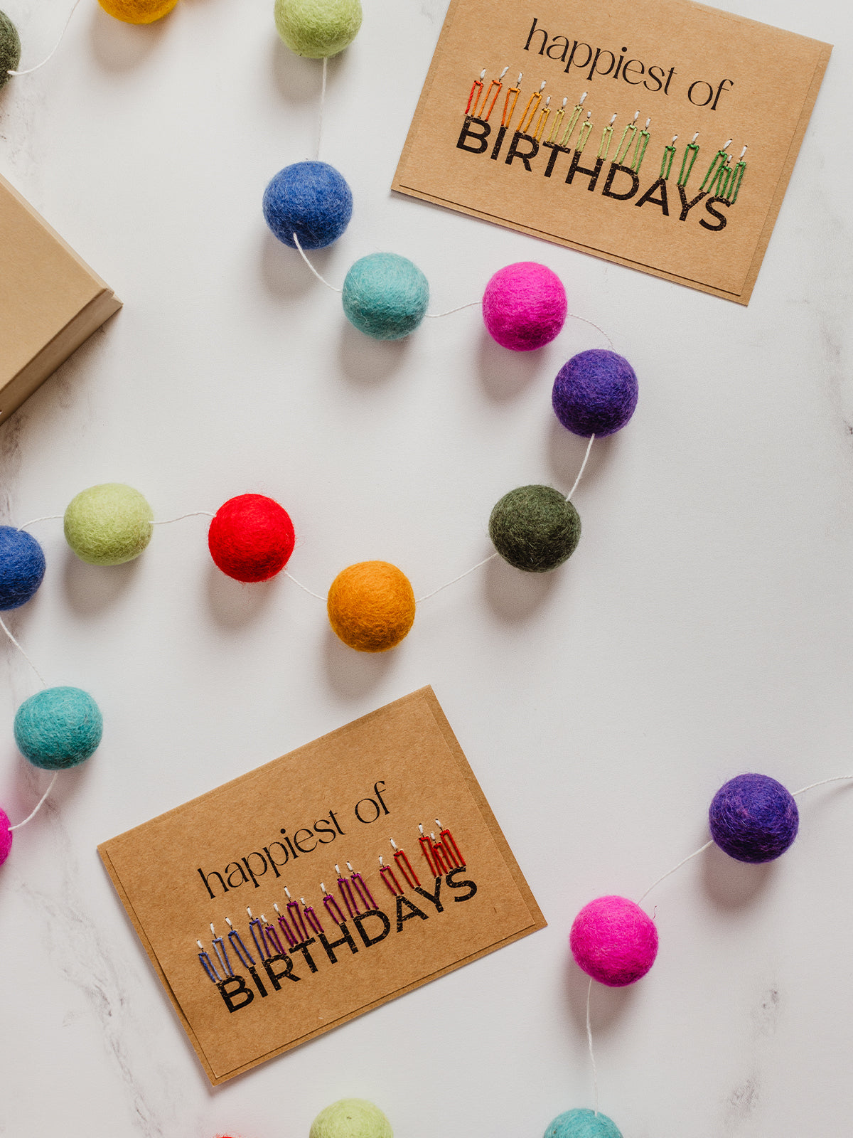 Flat lay of colorful felt ball garland with kraft paper cards reading “happiest of BIRTHDAYS” on a white marble background. Eco-friendly handmade party decor.