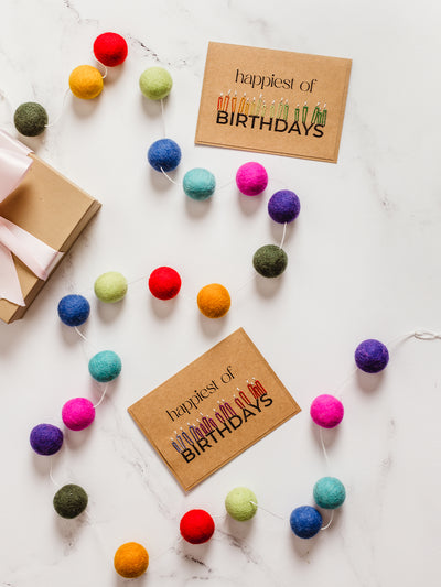 Sustainable birthday setup with a kraft gift box, pink ribbon, and vibrant felt ball garland with “happiest of BIRTHDAYS” cards on white marble. Eco-friendly handmade party decorations.
