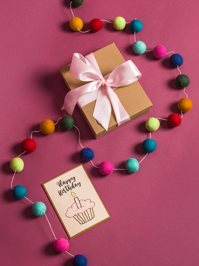 Eco-friendly birthday flat lay with a kraft gift box tied with a pink ribbon, colorful felt ball garland, and a “Happy Birthday” cupcake card on a deep pink background. Handmade wool party decoration and sustainable gift wrap idea.