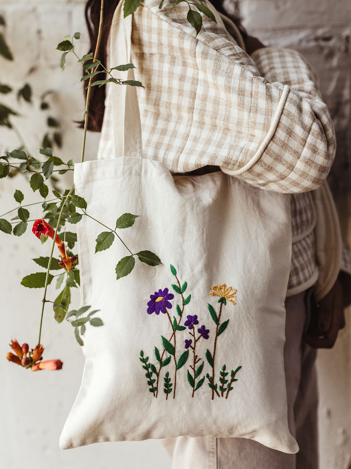 Close-up of a cream canvas tote bag with hand-embroidered floral design in purple and yellow flowers, styled with a neutral checkered jacket, ethical slow fashion bag.