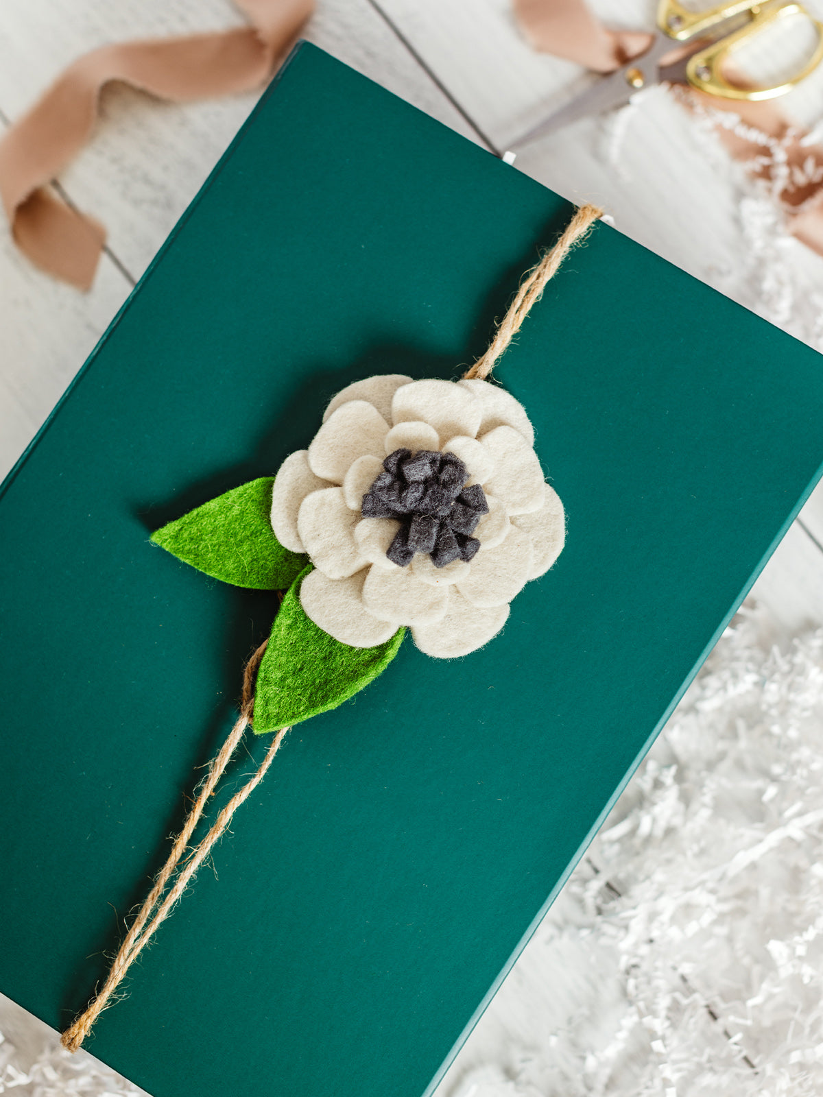 Handcrafted felt flower gift topper in cream, gray, and green tied with natural jute string on a dark green gift box — artisan-made sustainable gift wrapping accent.