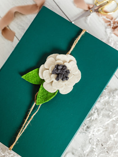 Handcrafted felt flower gift topper in cream, gray, and green tied with natural jute string on a dark green gift box — artisan-made sustainable gift wrapping accent.