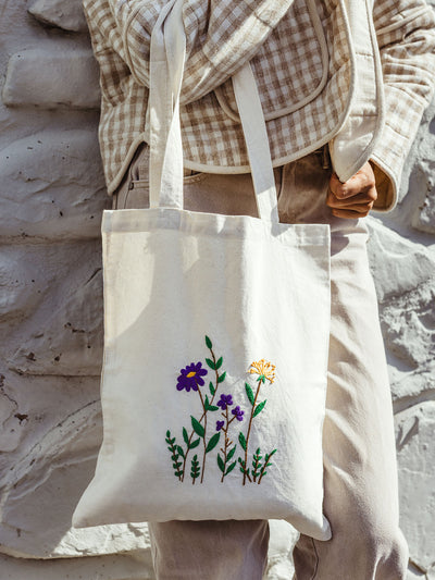 Handmade cotton tote bag with embroidered wildflowers in purple and yellow, eco-conscious reusable shopping bag styled with beige outfit, sustainable fashion accessory.