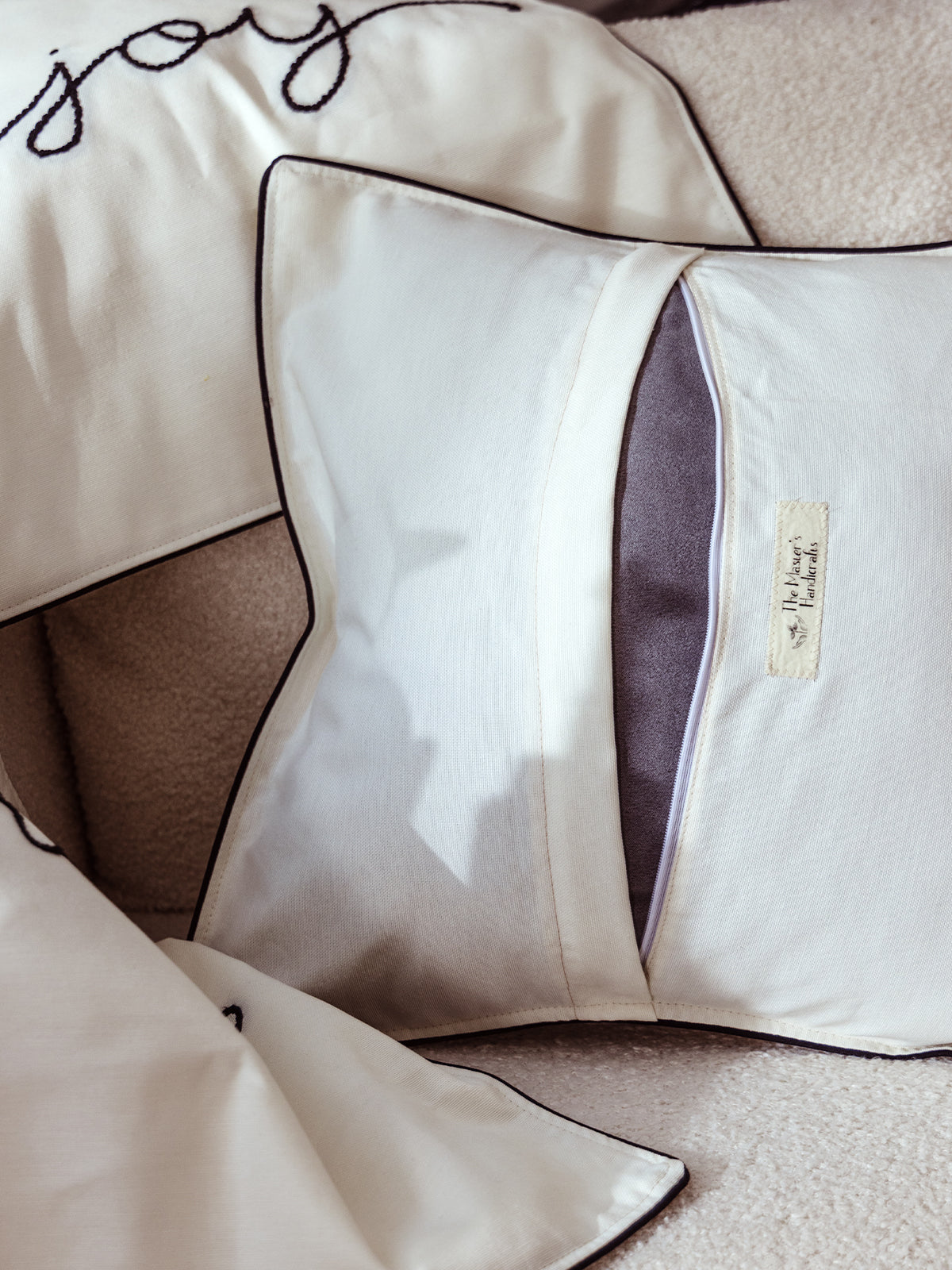 Back view of a handmade decorative pillow cover in cream fabric with dark piping, showing envelope closure and tag from Masters Handicrafts, styled on a cozy chair.