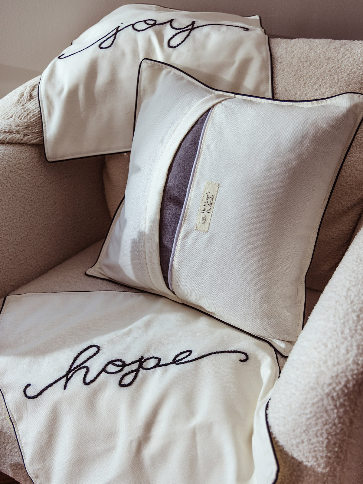 Set of cream throw pillows with black embroidered words “joy” and “hope,” displayed on a boucle armchair with a Masters Handicrafts pillow cover featuring envelope closure.