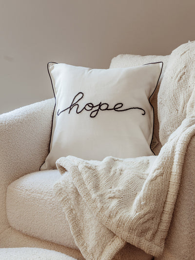 Neutral armchair styled with a cream decorative pillow embroidered with the word “hope” in cursive black thread, paired with a soft knitted throw blanket.
