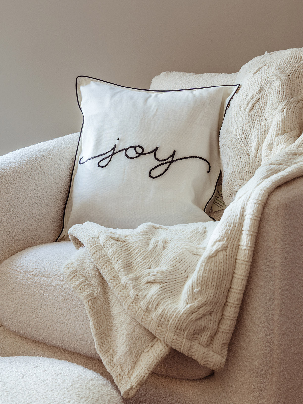 Minimalist holiday pillow with the word “joy” embroidered in black cursive on a cream cover with piped edges, styled on a boucle chair with a cozy knit blanket.