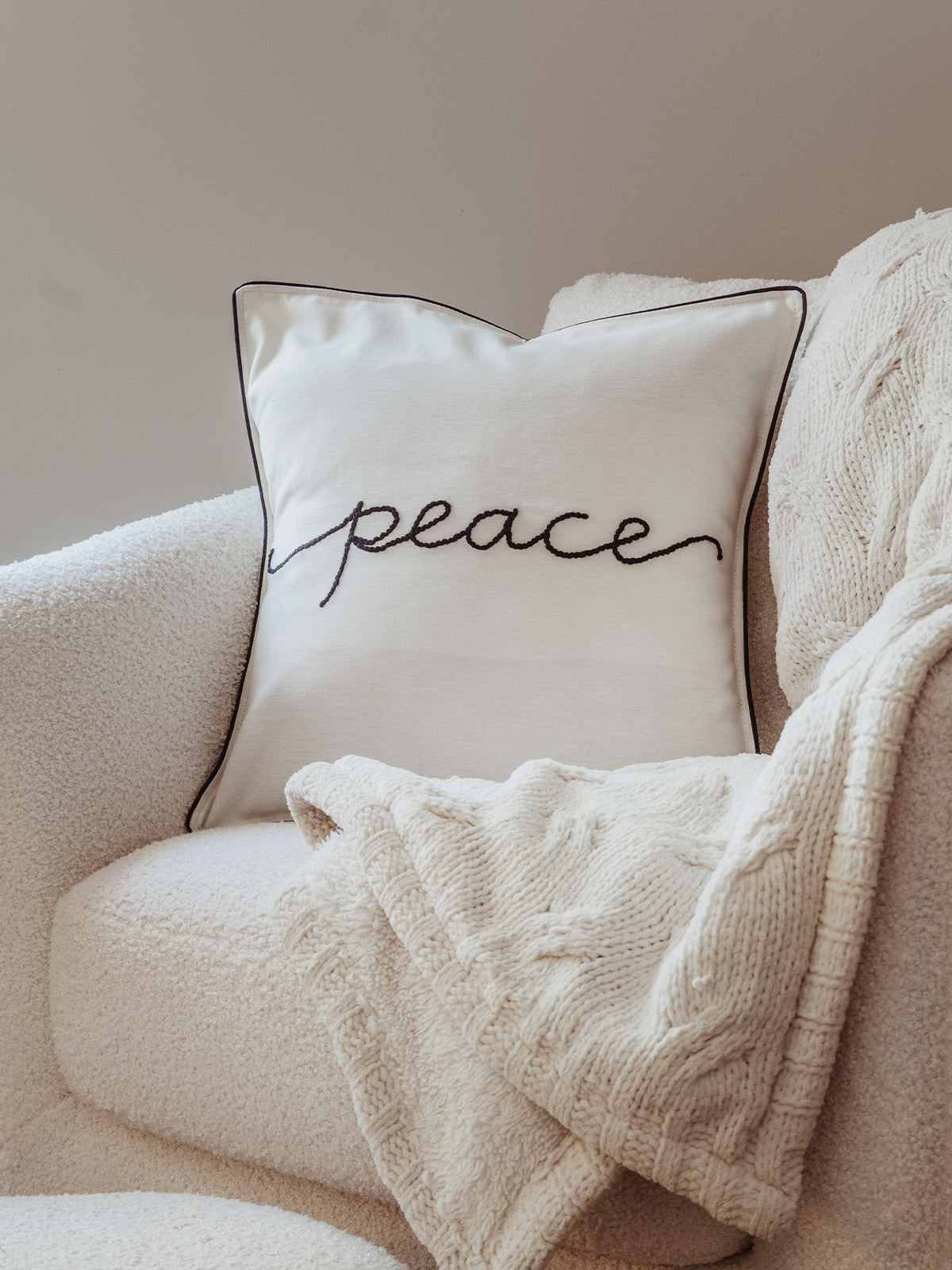 Cream throw pillow with black cursive embroidery spelling “peace,” styled on a boucle armchair with a neutral knit throw for a warm and inviting look.
