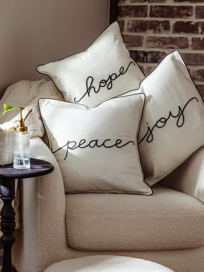Trio of decorative pillows with words “hope,” “peace,” and “joy” embroidered in black thread on cream fabric, arranged on a cozy armchair with a knit throw blanket.
