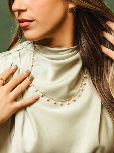 Close-up of a woman wearing a gold chain necklace with pearl drop accents, styled with a soft sage satin blouse and gold bow-shaped earrings.