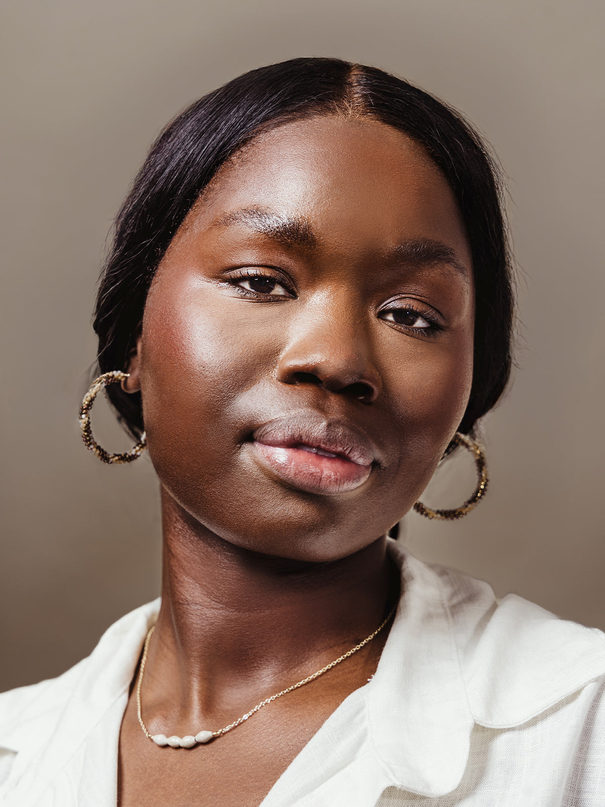 Close-up portrait of a woman wearing large gold and beaded hoop earrings and a delicate pearl and gold necklace, styled with a white top against a neutral background.