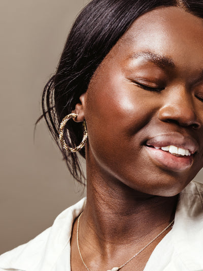 Detail shot of a woman smiling with her eyes closed, showcasing large handcrafted hoop earrings made of gold and multicolored beads.