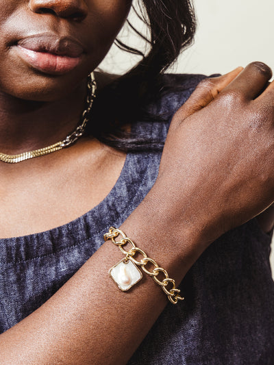 Close-up of a woman wearing a chunky gold chain bracelet with a square pearl charm, styled with a dark denim sleeveless top and a layered gold necklace.