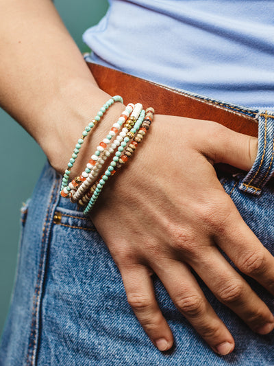 Flat lay of vibrant handmade beaded bracelets in turquoise, coral, white, and earth tones styled around halved oranges on a pastel blue surface, creating a fresh and colorful aesthetic.