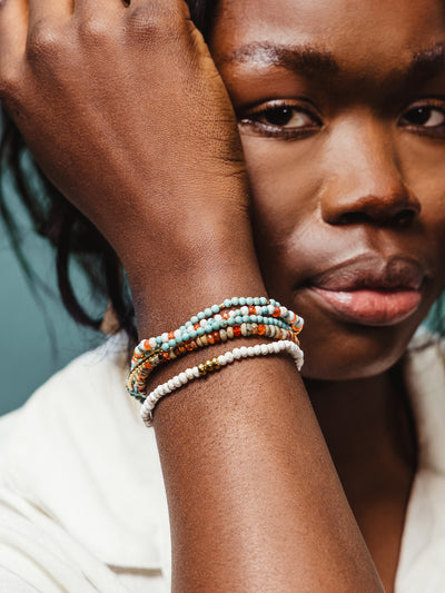 Close-up of a woman with her arm raised, wearing a stack of multicolored beaded bracelets featuring turquoise, white, coral, and gold accents, styled with a neutral blouse.