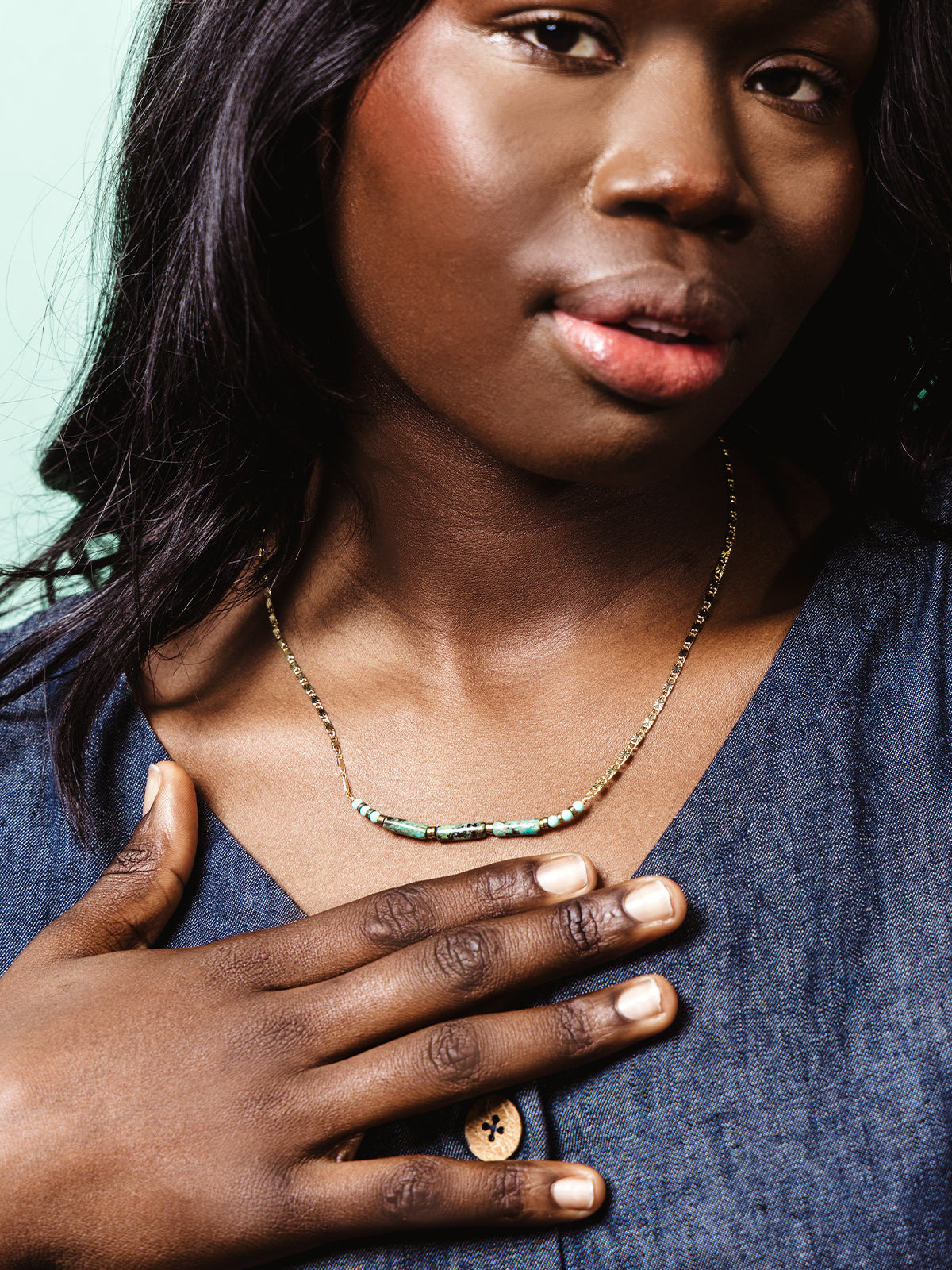 Close-up of a woman wearing a delicate gold chain necklace with turquoise and green stone beads, styled with a denim top and her hand gently resting on her chest.