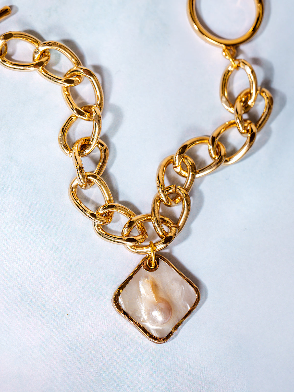 Close-up of a gold link bracelet with a unique square-shaped pearl charm, displayed on a soft white background with natural light reflecting off the polished metal.