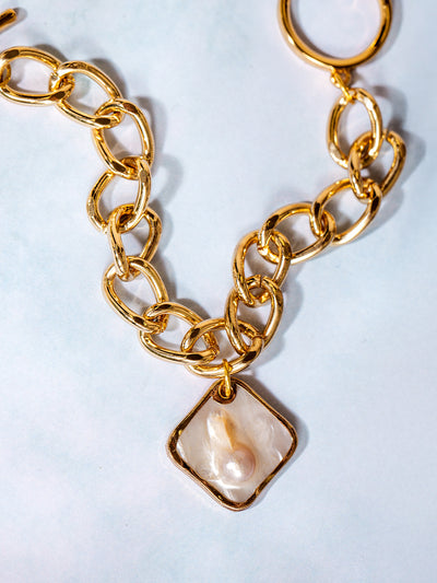 Close-up of a gold link bracelet with a unique square-shaped pearl charm, displayed on a soft white background with natural light reflecting off the polished metal.