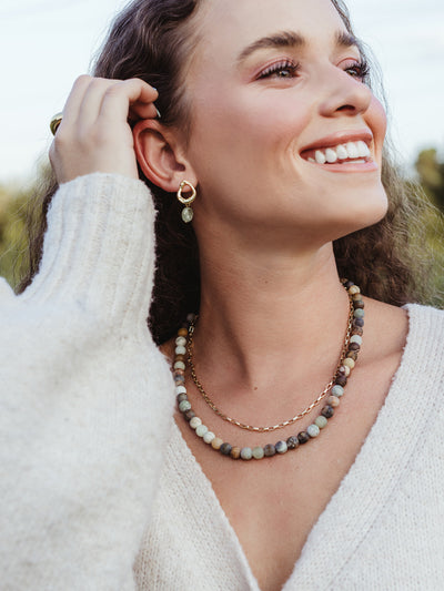 Smiling woman in cream knit sweater wearing gold gemstone drop earrings and layered necklaces with natural stone beads and gold chain.