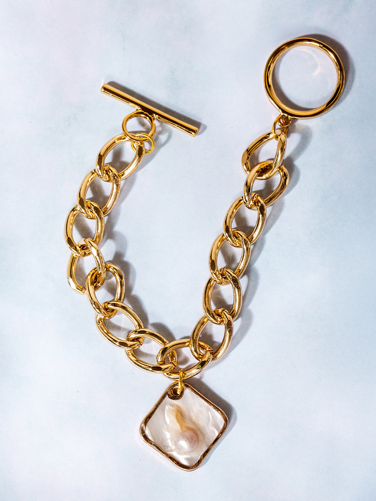Flat lay of a gold chain toggle bracelet featuring a square pearl pendant, arranged in a semi-circle on a light background with a minimal and elegant aesthetic.