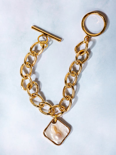 Flat lay of a gold chain toggle bracelet featuring a square pearl pendant, arranged in a semi-circle on a light background with a minimal and elegant aesthetic.