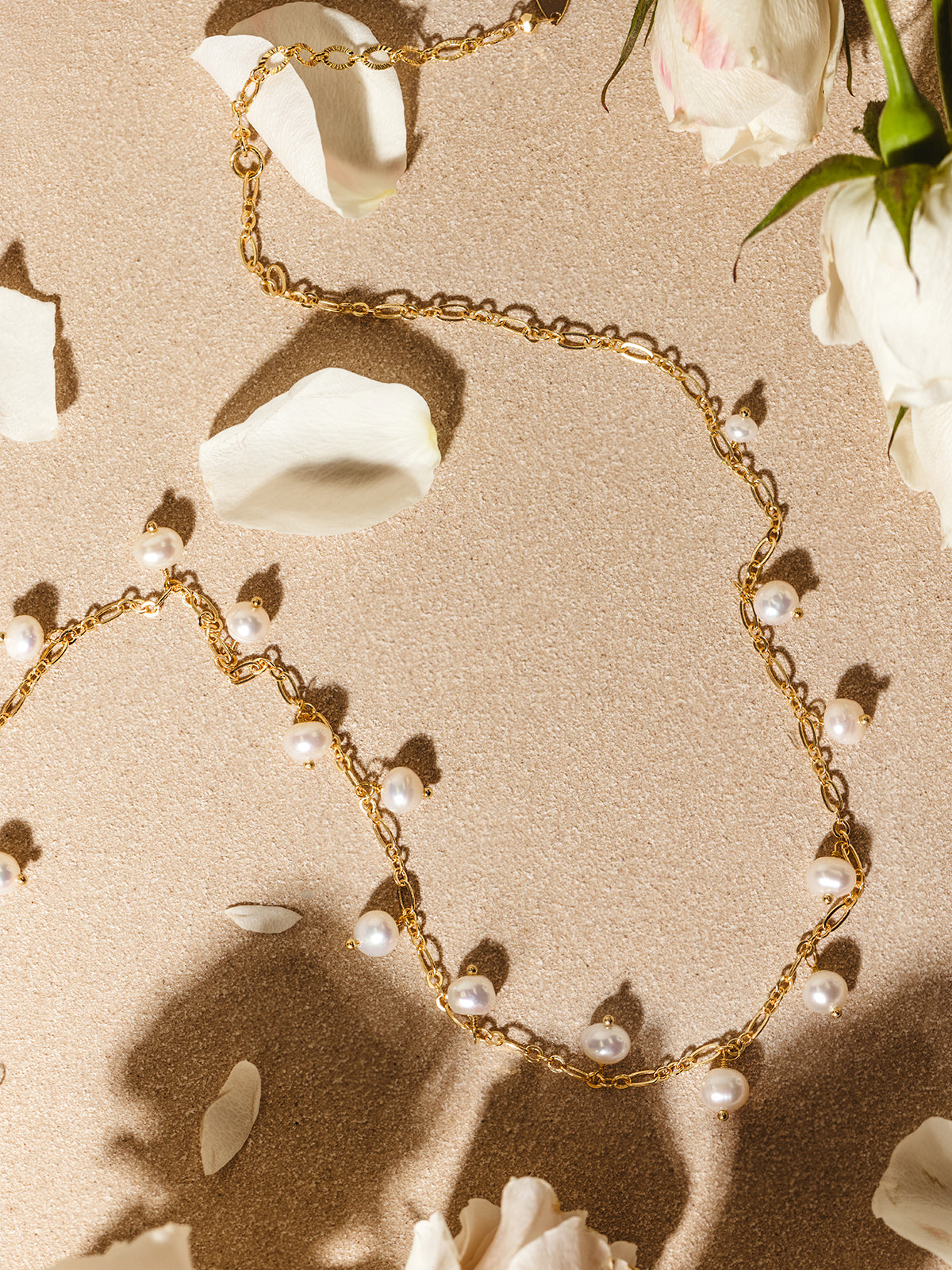 Flat lay of a gold chain necklace adorned with dangling white freshwater pearls, styled among white rose petals on a softly lit neutral background.