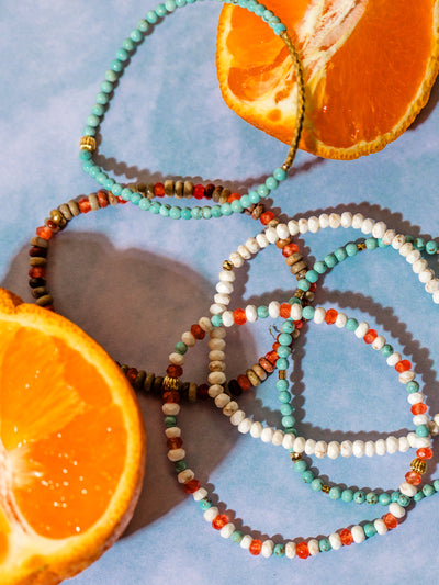 Flat lay of vibrant handmade beaded bracelets in turquoise, coral, white, and earth tones styled around halved oranges on a pastel blue surface, creating a fresh and colorful aesthetic.