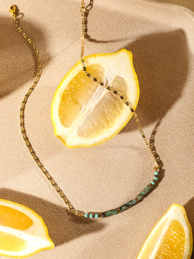 Flat lay of a gold chain necklace with turquoise and green beads, styled alongside sliced lemons on a sunlit neutral surface for a bright, summery vibe.