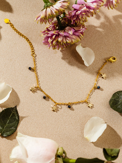 Flat lay of a gold chain bracelet with star-shaped charms and dark blue beads, styled among white rose petals and purple flowers on a soft neutral surface.