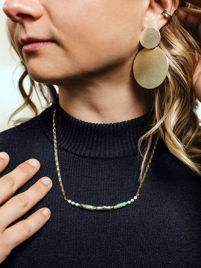 Side view of a woman wearing a gold beaded necklace with turquoise and green accents, styled with a navy ribbed turtleneck and large matte gold statement earrings.