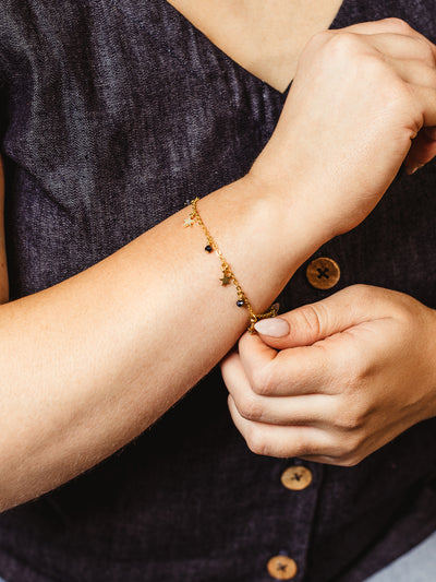 Close-up of a woman adjusting a delicate gold chain bracelet adorned with tiny star charms and dark gemstone beads, styled with a sleeveless indigo top.