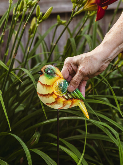 Hand placing a painted bird garden stake among tall green leaves and blooming daylilies in a garden bed.