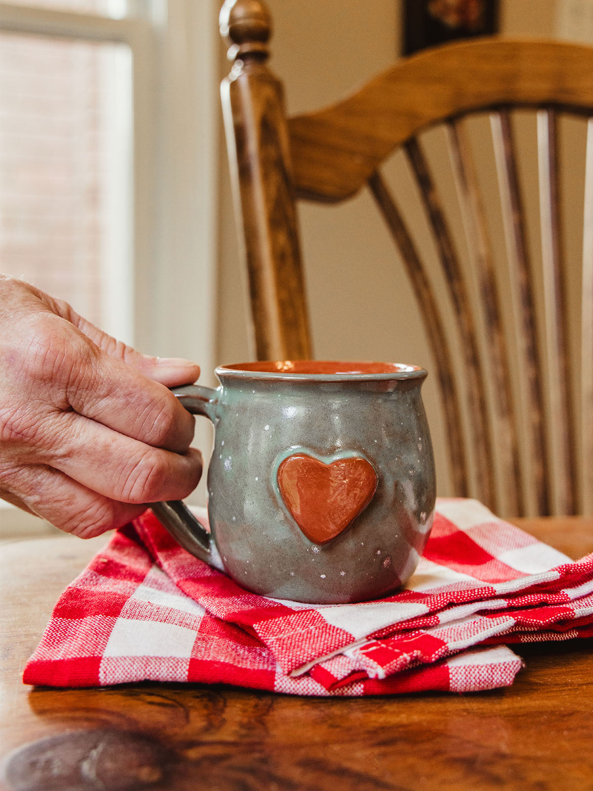 A hand holding a handcrafted ceramic mug with a red heart detail on the front, resting on a red and white checkered cloth atop a wooden table, with a wooden chair and window in the background.