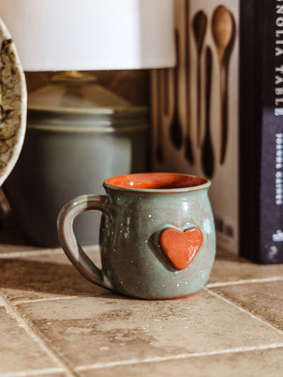 Handcrafted ceramic mug with a glossy green glaze and a raised red heart on the front, featuring a rounded shape and curved handle. Styled on a tiled kitchen counter next to a cookbook and a ceramic lamp.