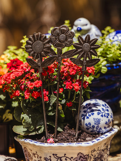 A trio of metal flower garden stakes styled in a floral ceramic planter surrounded by bright red flowers and a blue-and-white decorative sphere in the background.