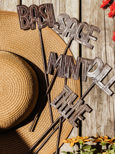 A flat lay of metal herb garden markers labeled Basil, Sage, Mint, Dill, and Thyme, styled on top of a woven straw hat on a wooden surface.