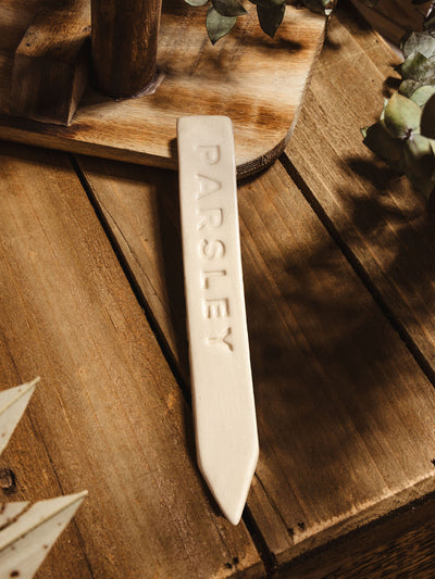 Ceramic garden marker "parsley" lying on a wooden surface with some greenery. 