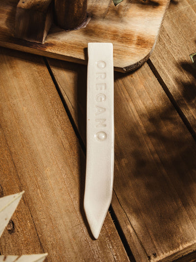 Ceramic garden marker "oregano" lying on a wooden surface with some greenery. 