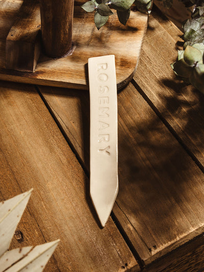 Ceramic garden marker "rosemary" lying on a wooden surface with some greenery. 