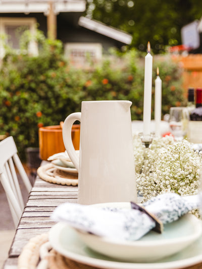 Off white ceramic pitcher placed on outdoor dinning table in backyard setting.