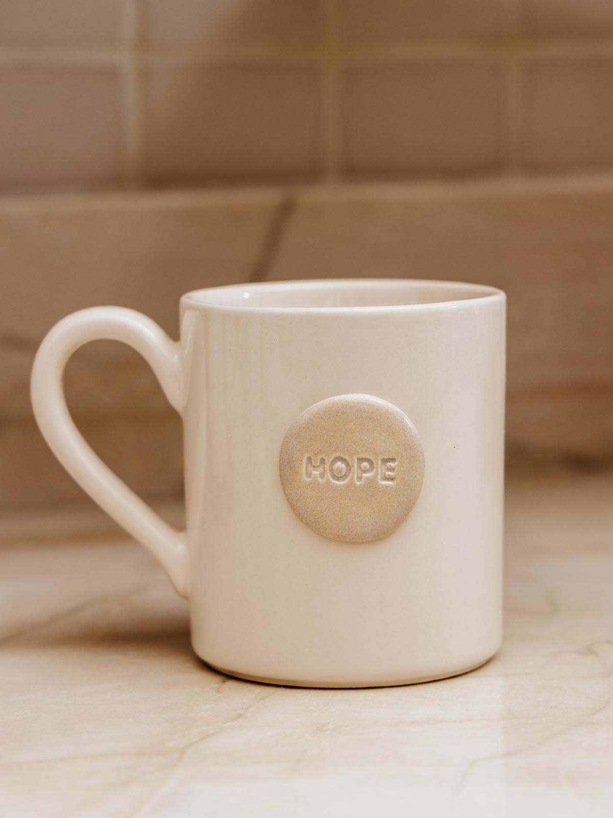 Ceramic white mug with circular ceramic piece that says "hope" sitting on white counter top.