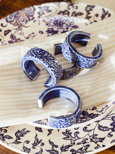 Overhead view of four blue ceramic napkin rings with floral motifs resting on a striped cloth napkin and stacked blue and white plates, capturing a refined and artisan tabletop look.