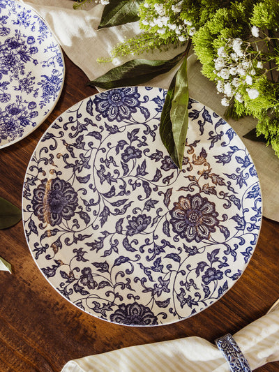 Flat lay of a large blue and white floral charger plate surrounded by greenery and baby's breath, styled on a dark wood table.