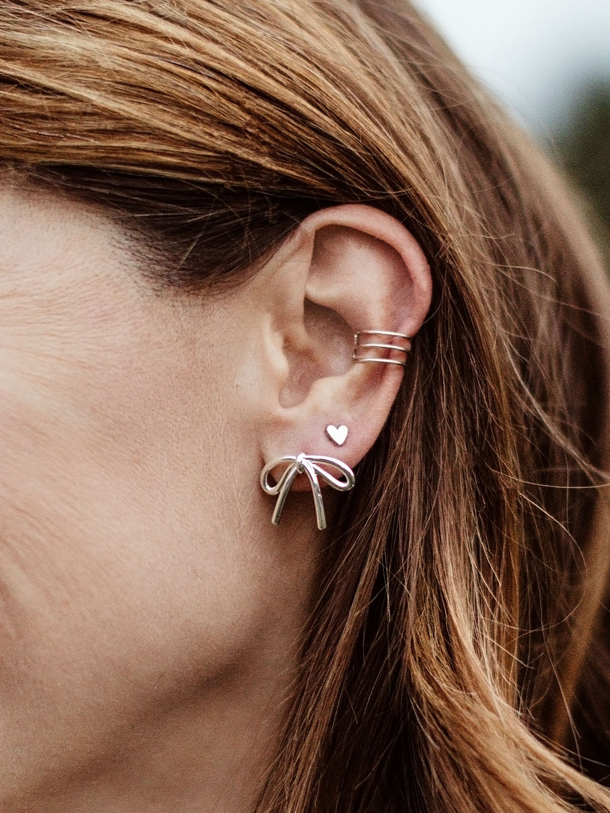 Close-up of silver bow stud earrings paired with minimalist ear cuff and tiny heart stud, modern ethical jewelry set.