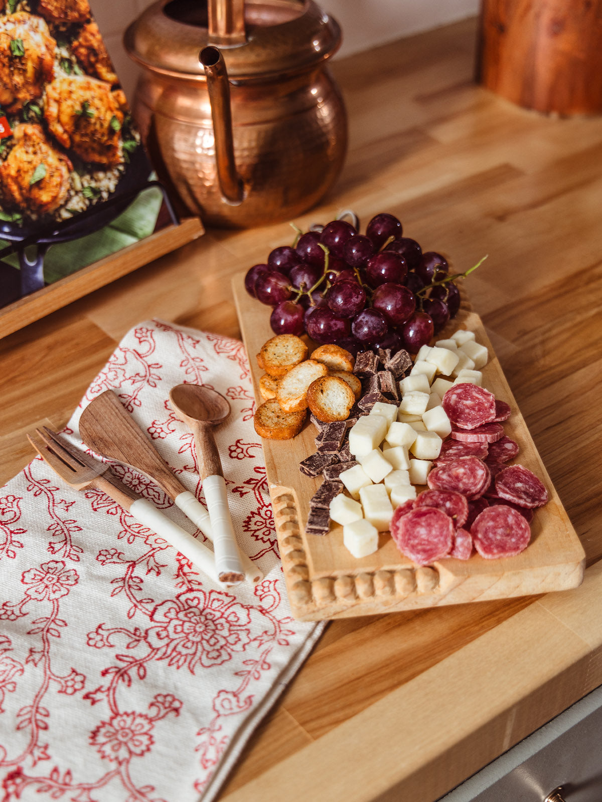 Artisan kitchen gift bundle featuring a hand-carved wooden serving board with grapes, cheese, salami, crackers, and chocolate, paired with red floral block print tea towel and wooden utensils, ethically made home entertaining set.