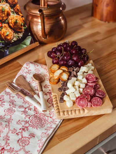 Artisan kitchen gift bundle featuring a hand-carved wooden serving board with grapes, cheese, salami, crackers, and chocolate, paired with red floral block print tea towel and wooden utensils, ethically made home entertaining set.