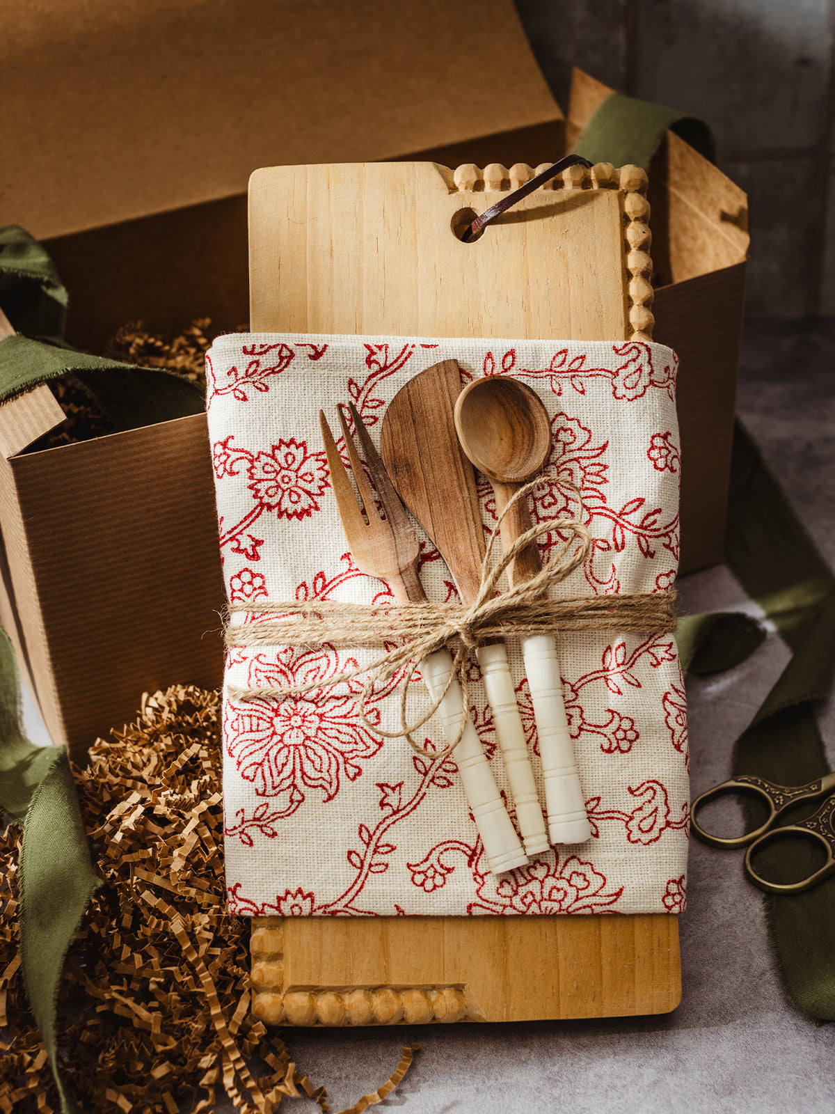 Eco-friendly kitchen gift bundle featuring a wooden board, red floral cloth, and handmade wooden utensils in rustic holiday packaging