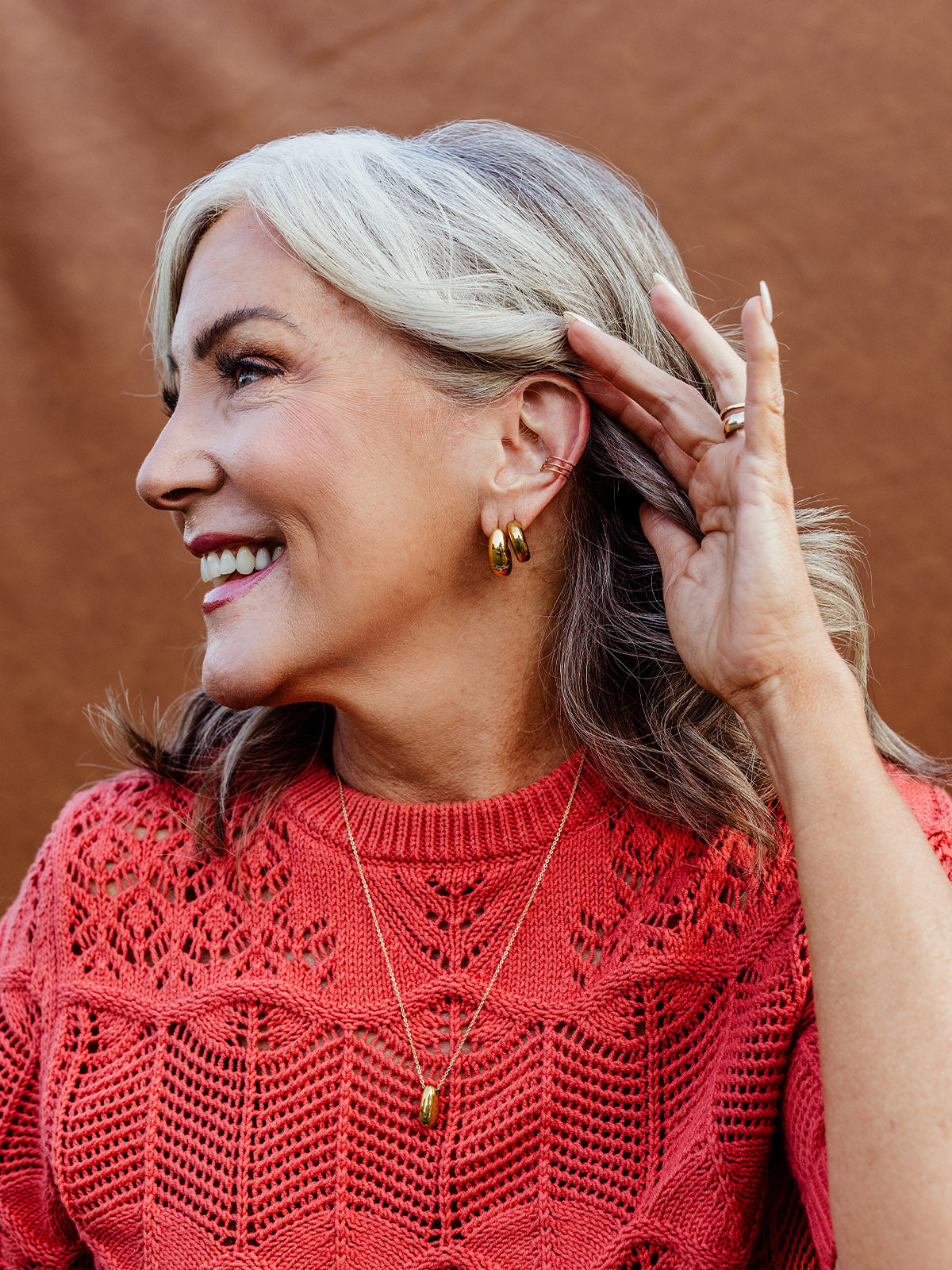 Smiling woman wearing gold huggie hoop earrings and matching pendant necklace, styled with a coral knit sweater, showcasing the Huggie Hoop Bundle — ethically made minimalist jewelry for everyday wear.