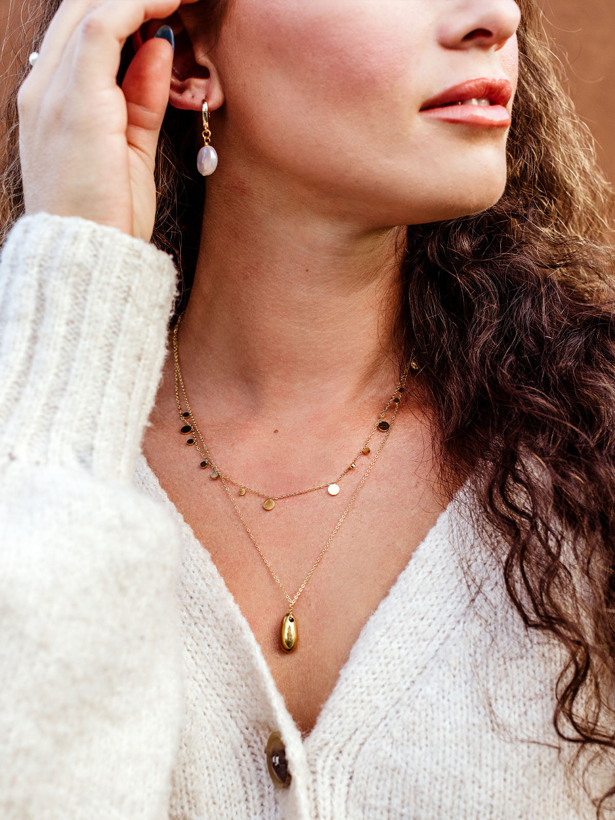Woman wearing gold layered necklaces with disc charm chain and teardrop pendant, styled with pearl drop earrings and cozy cream sweater, minimalist everyday jewelry set for women.
