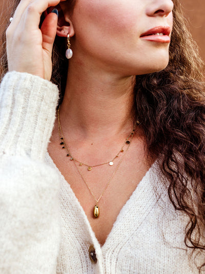 Woman wearing gold layered necklaces with disc charm chain and teardrop pendant, styled with pearl drop earrings and cozy cream sweater, minimalist everyday jewelry set for women.